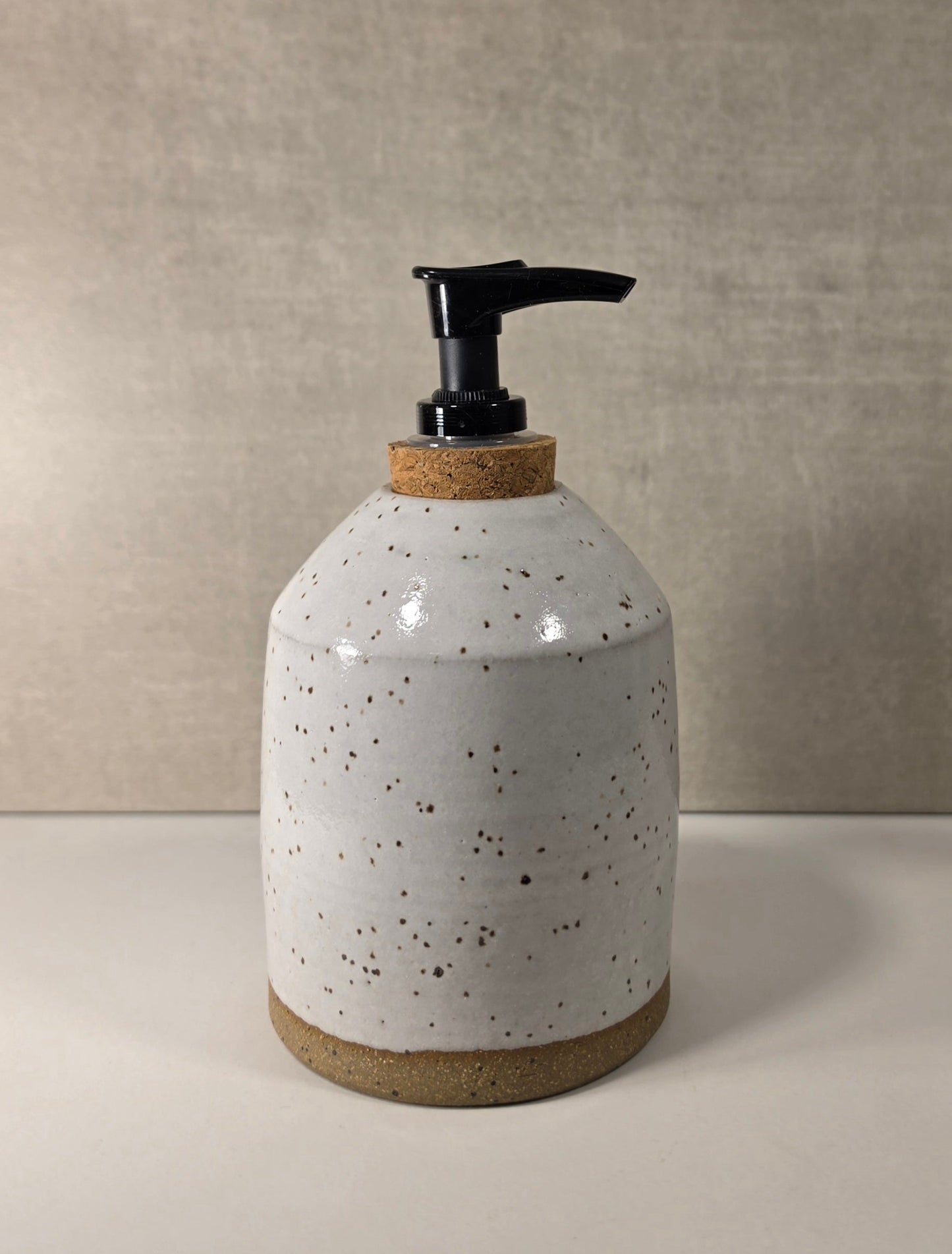 A ceramic soap dispenser with a speckled off-white glaze, a natural cork neck, and a black plastic pump, set against a neutral textured beige background.