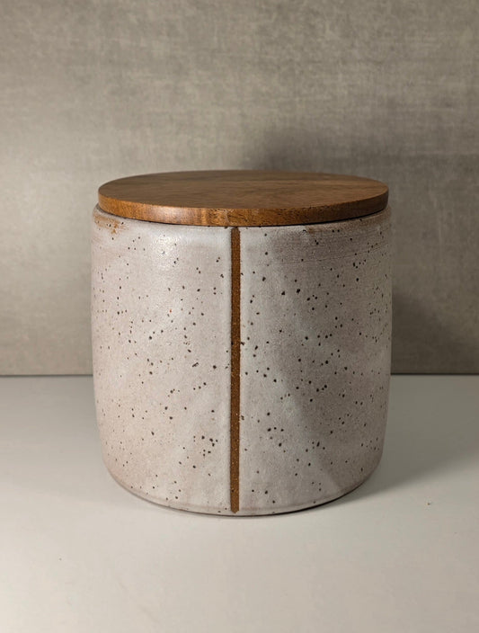A cylindrical ceramic storage jar with a speckled light gray and white glaze, featuring a vertical brown wood grain stripe down the center and a matching round wooden lid, set against a neutral gray textured background.