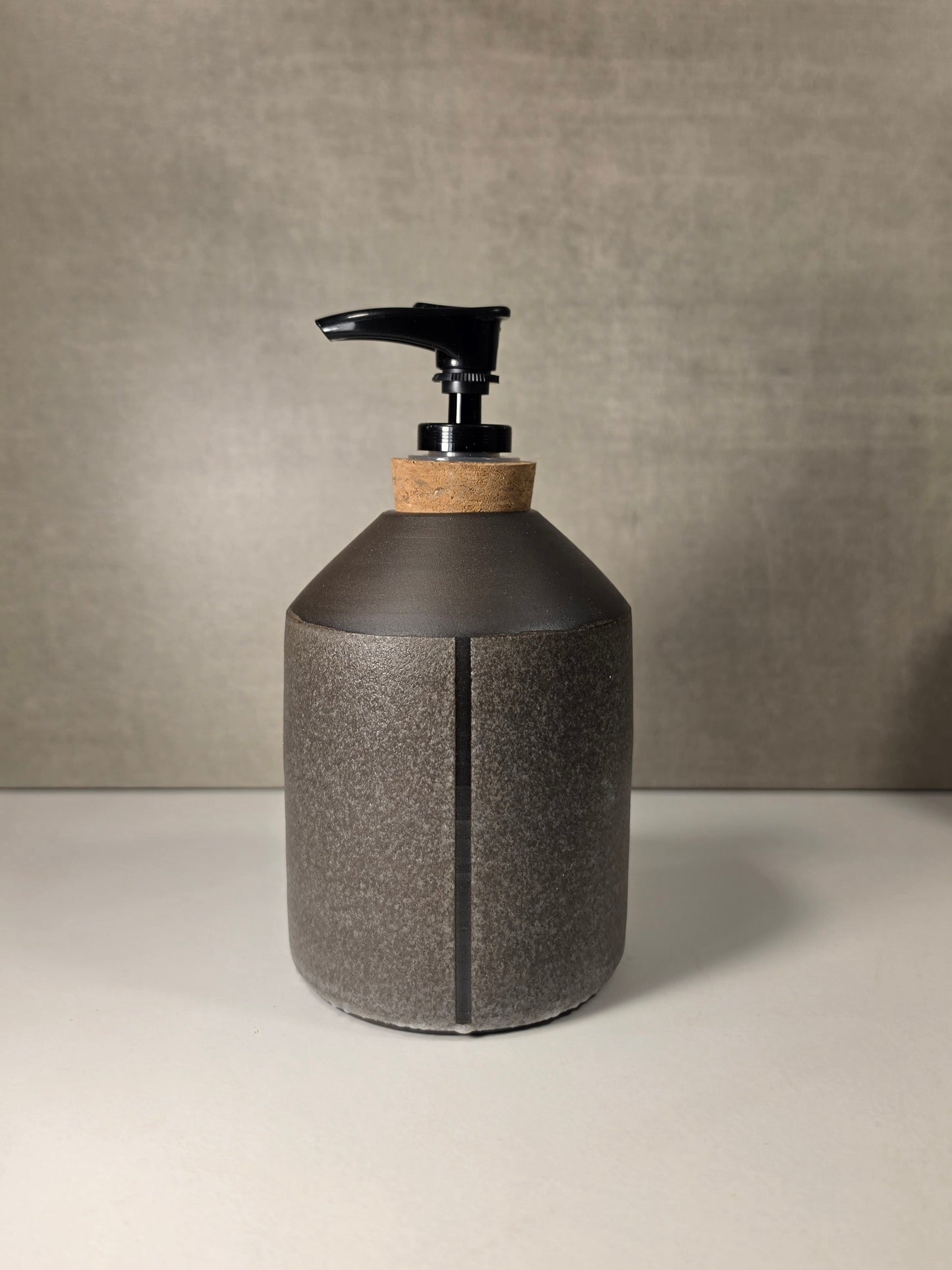 A dark gray textured dispenser bottle with a cork neck and a black pump dispenser, set against a neutral gray background.