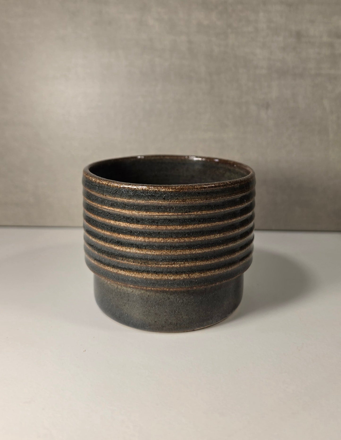 A dark green ceramic cup with horizontal ridges which the glaze breaks over, set against a neutral gray background.