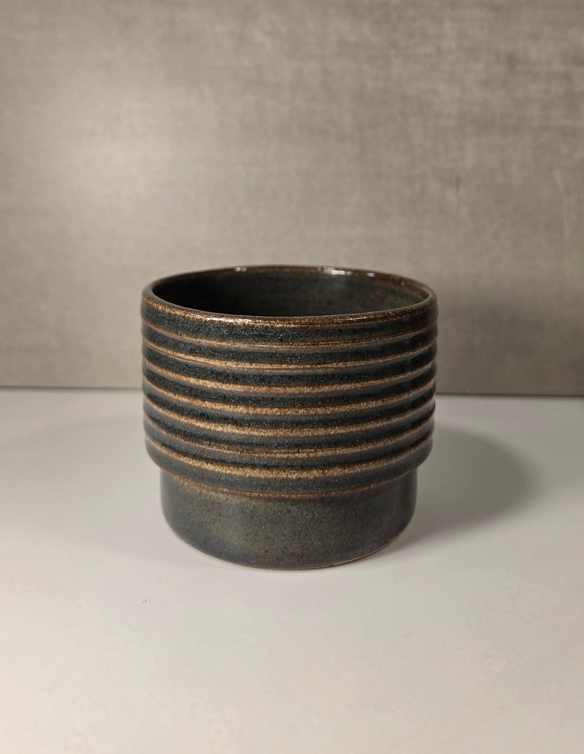 A dark green ceramic cup with horizontal ridges which the glaze breaks over, set against a neutral gray background.