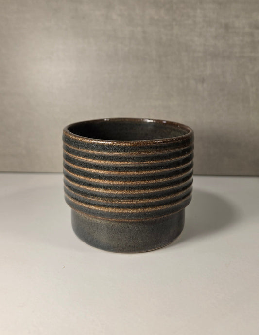 A dark green ceramic cup with horizontal ridges which the glaze breaks over, set against a neutral gray background.