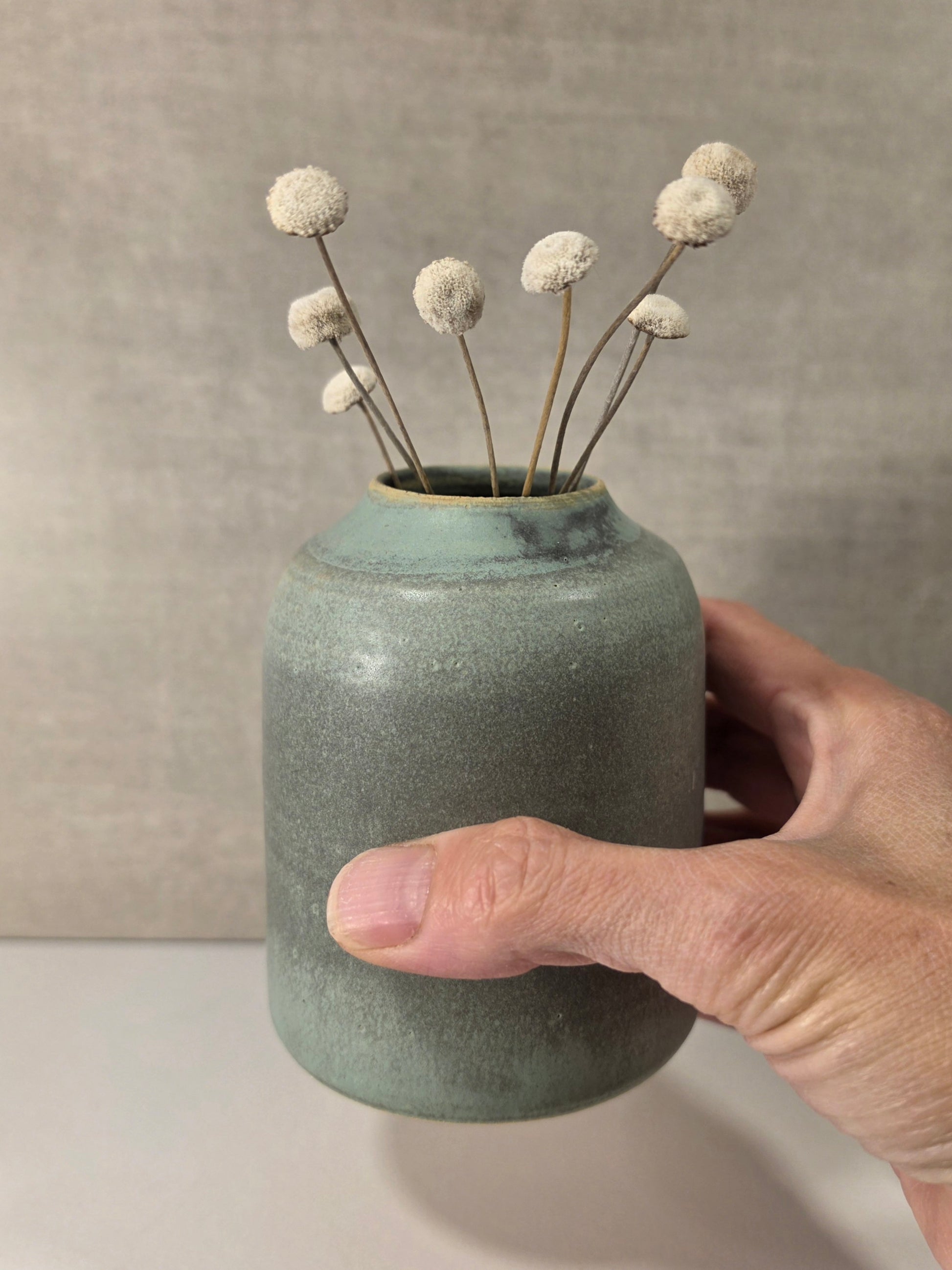 A hand holding a small round ceramic vase with a muted sage green glaze, decorated with dried white globe flowers on thin stems, set against a neutral textured gray background.