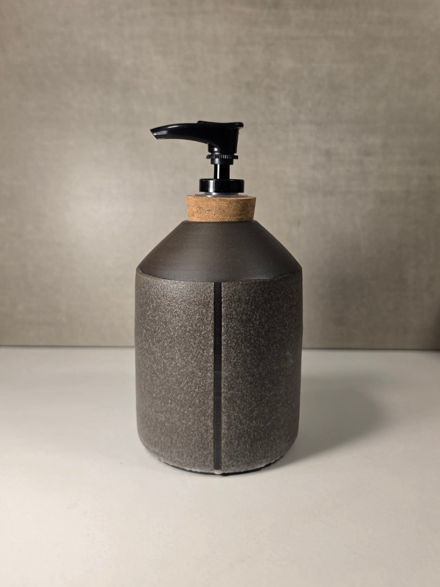 A dark gray textured ceramic pump bottle with a cork accent and black dispenser, set against a neutral gray background.