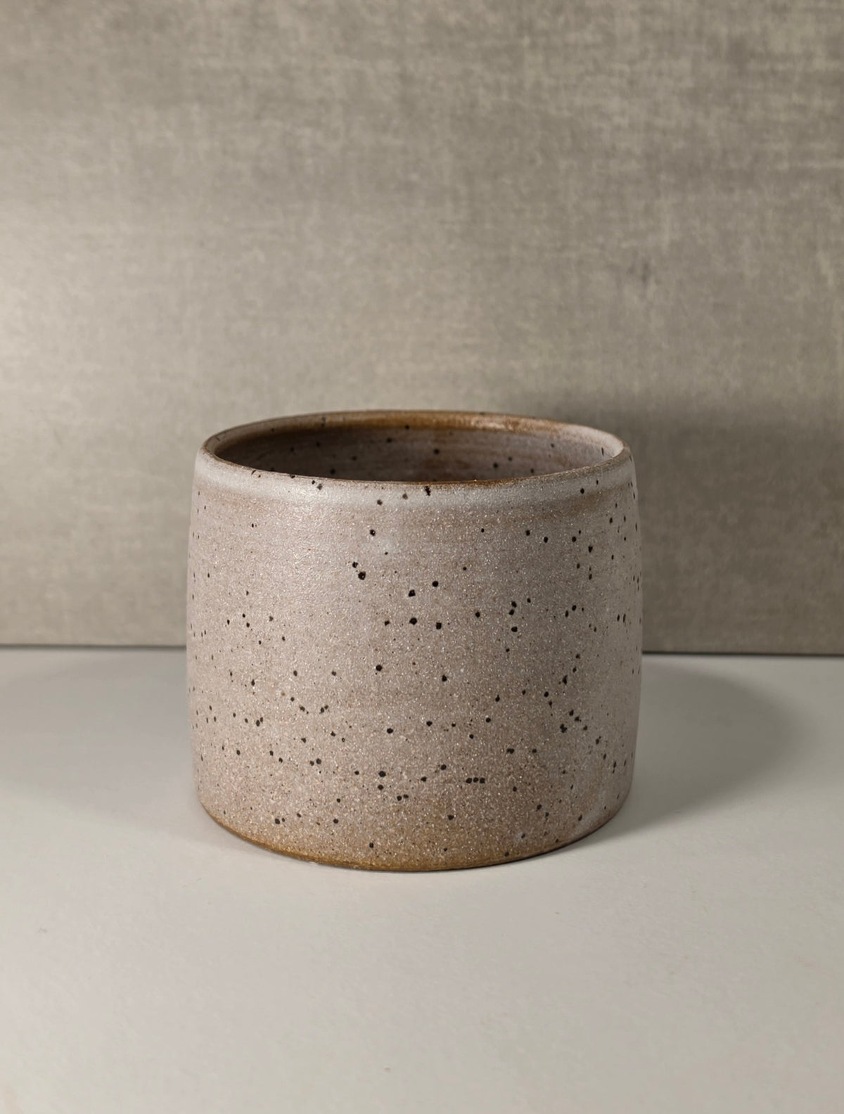 A small cylindrical ceramic planter with a speckled, textured matte finish in a light taupe-beige color, set against a neutral gray background.
