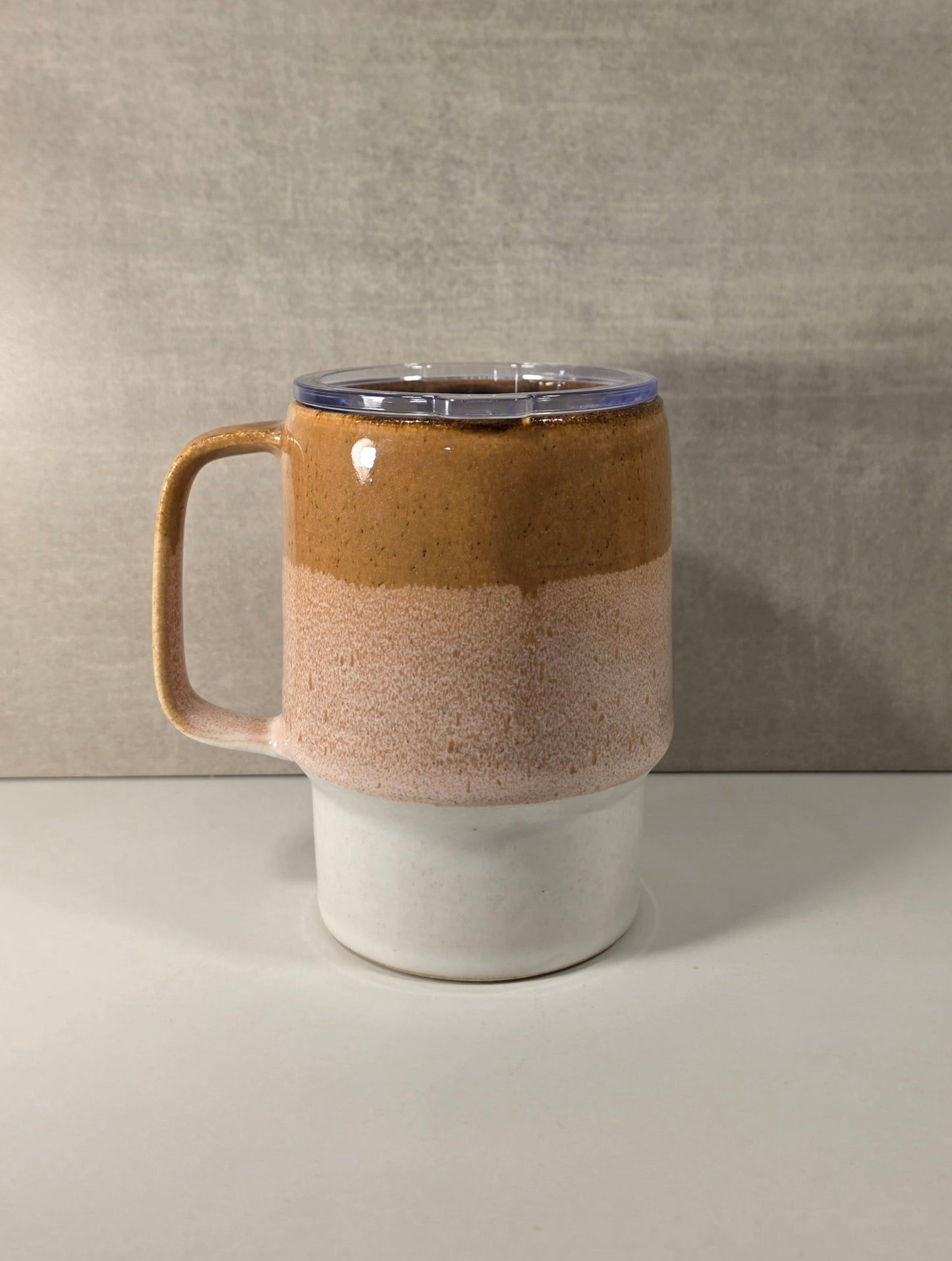 A ceramic travel mug with a handle, featuring a three-tone glaze design: a dark brown top section, a speckled light brown and pink middle section, and a solid white bottom section. It is shown with a clear plastic lid on a neutral gray background.