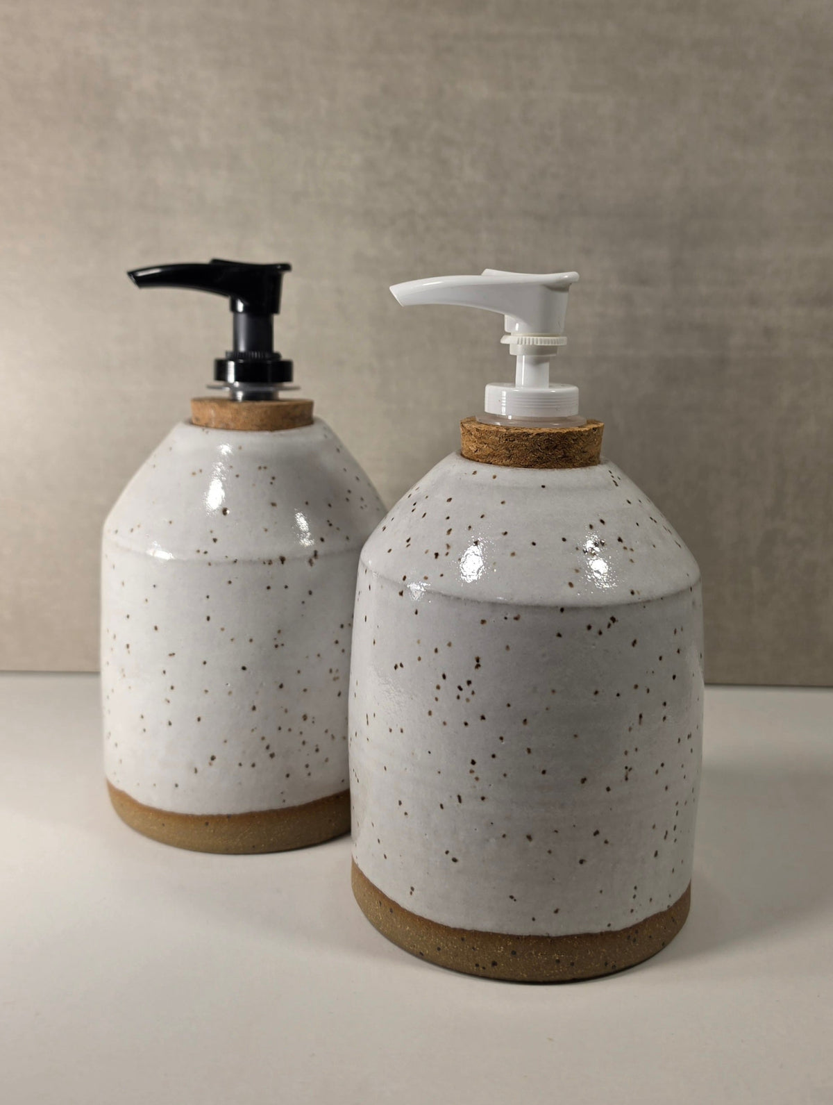 Two ceramic soap dispensers with speckled off-white glaze, cork accents, and pump dispensers (one black, one white) on a neutral beige background.