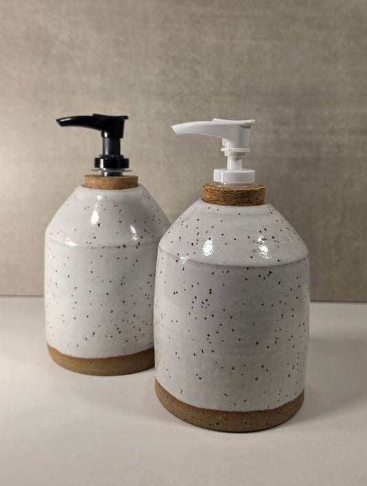 Two ceramic soap dispensers with speckled off-white glaze, cork accents, and pump dispensers (one black, one white) on a neutral beige background.