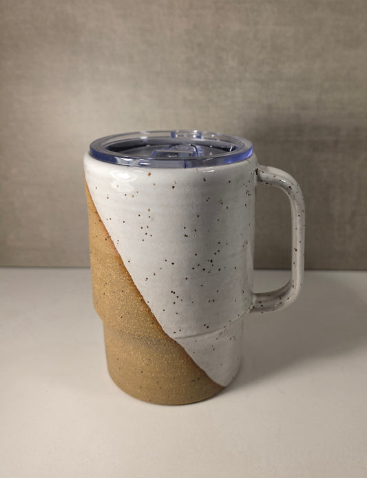 A ceramic travel tumbler with a diagonal split design, featuring a speckled off-white glaze on the top half and a textured tan clay finish on the bottom half. It has a clear plastic lid with a blue tint and a sturdy handle, set against a neutral beige background.