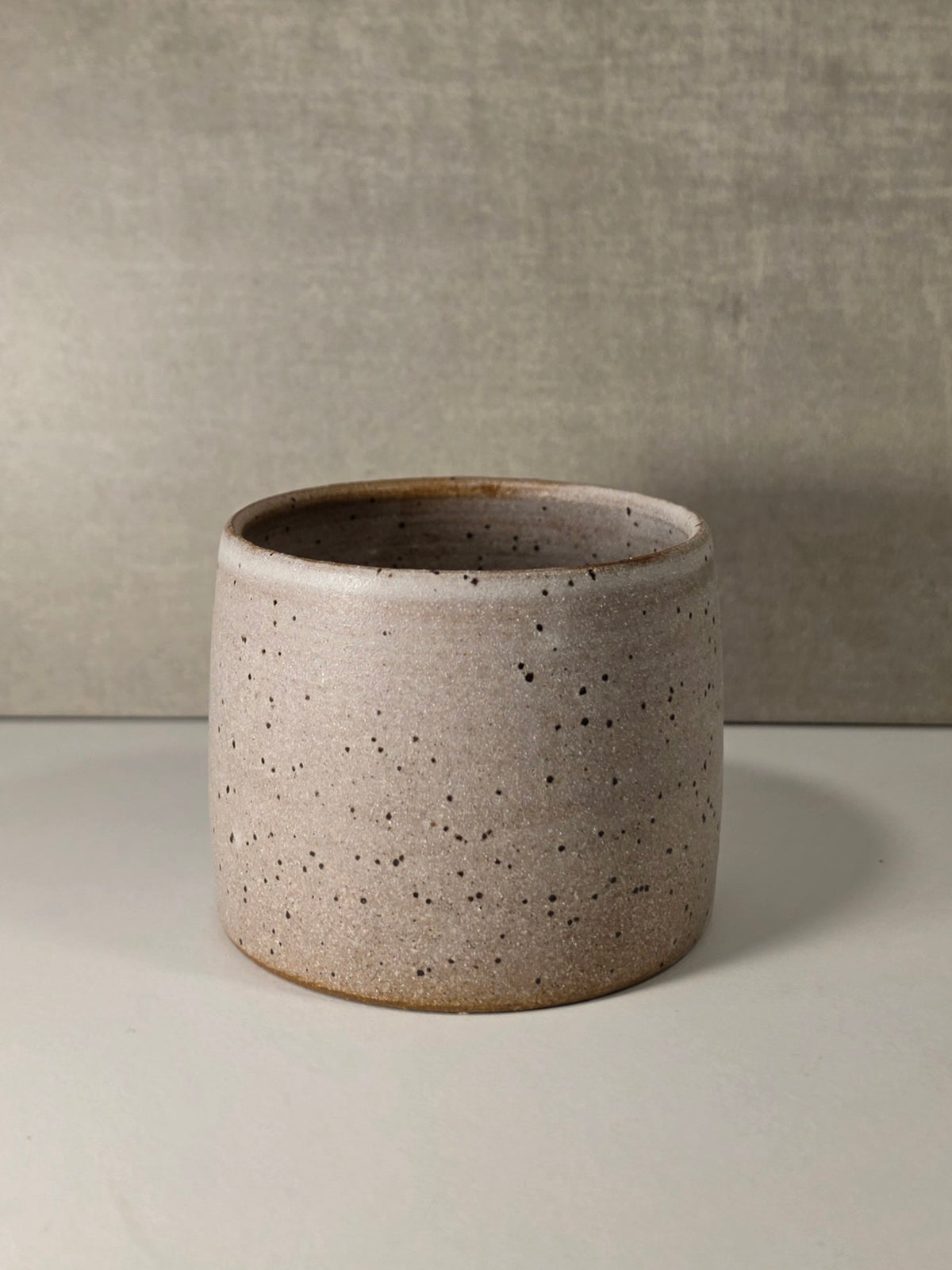 A small cylindrical ceramic planter with a speckled, matte stoneware finish in a light beige-gray color, set against a neutral textured gray background.