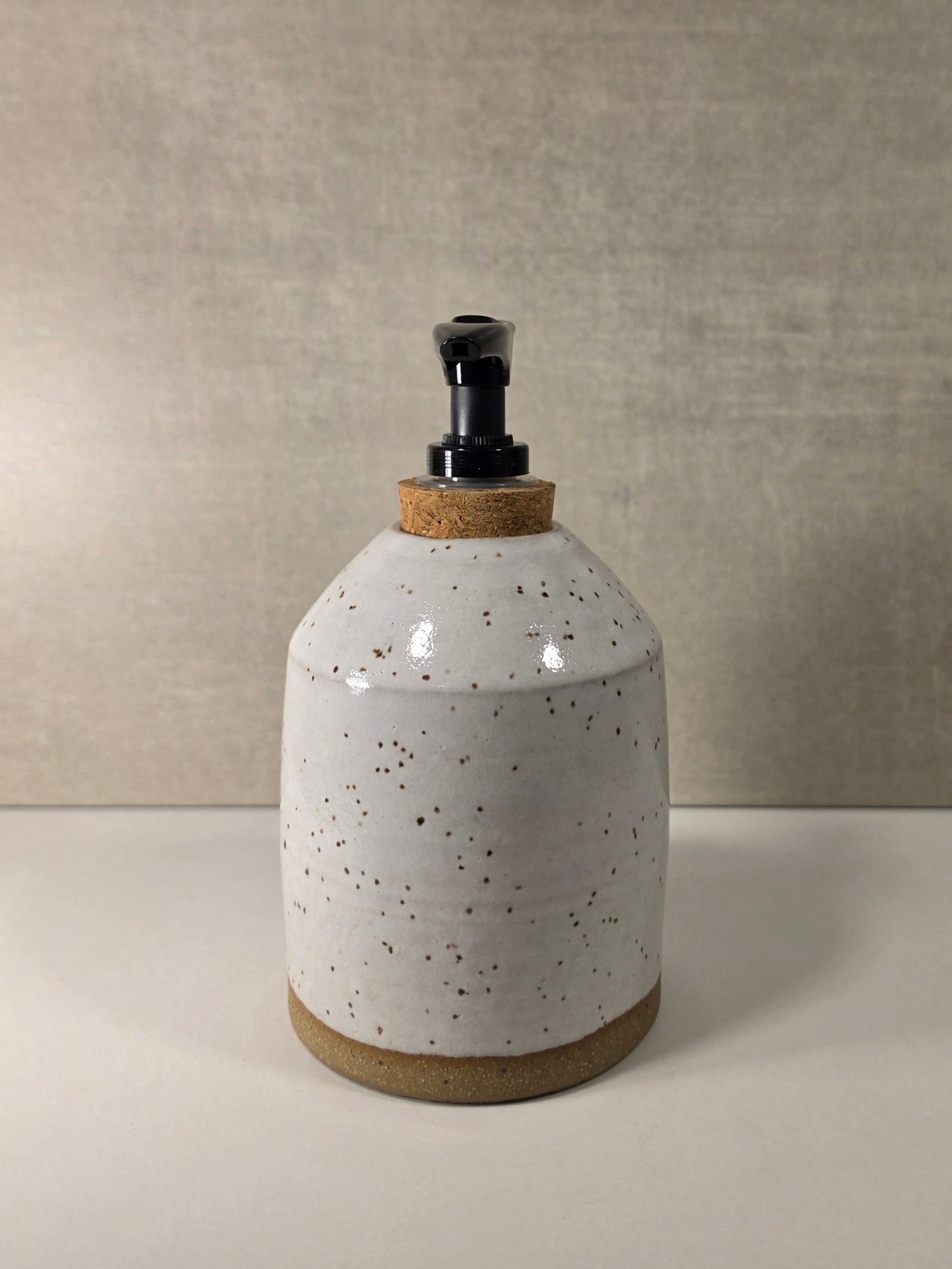 A handmade ceramic soap dispenser bottle with a speckled white glaze, a natural cork stopper, and a black plastic pump, set against a neutral textured beige background.