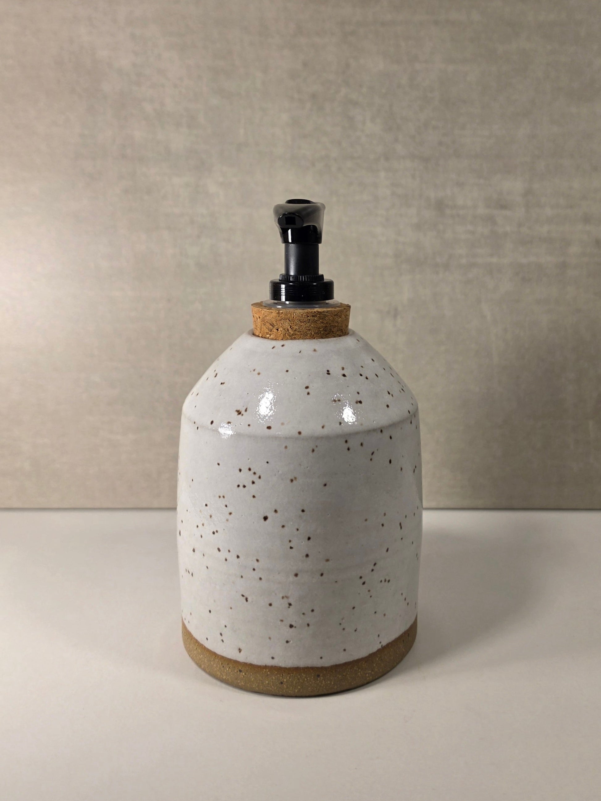 A handmade ceramic soap dispenser bottle with a speckled white glaze, a natural cork stopper, and a black plastic pump, set against a neutral textured beige background.