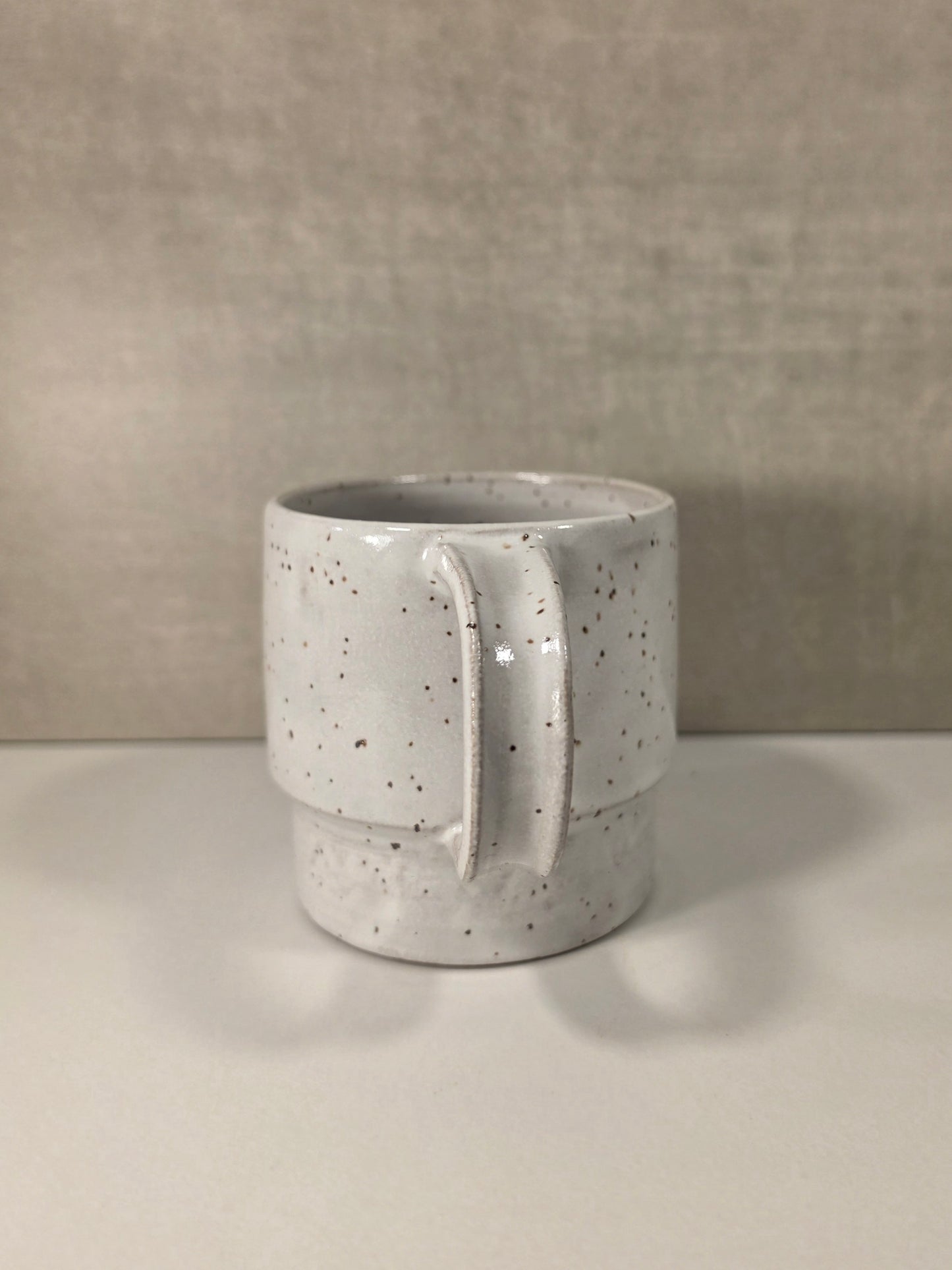 A handmade ceramic mug with a textured, speckled off-white glaze and a stacked tiered design, set against a neutral beige background.