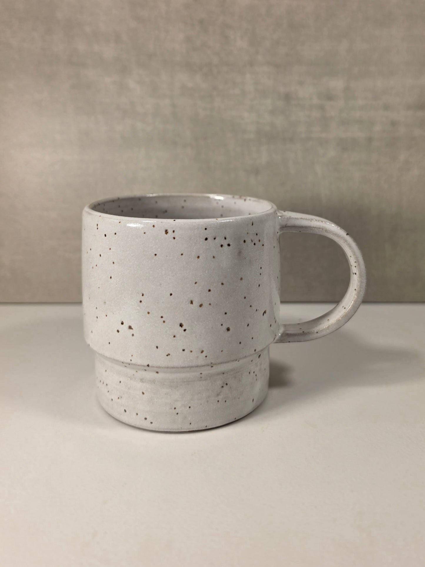 A handmade ceramic mug with a textured, speckled off-white glaze and a thick handle, set against a neutral gray background.