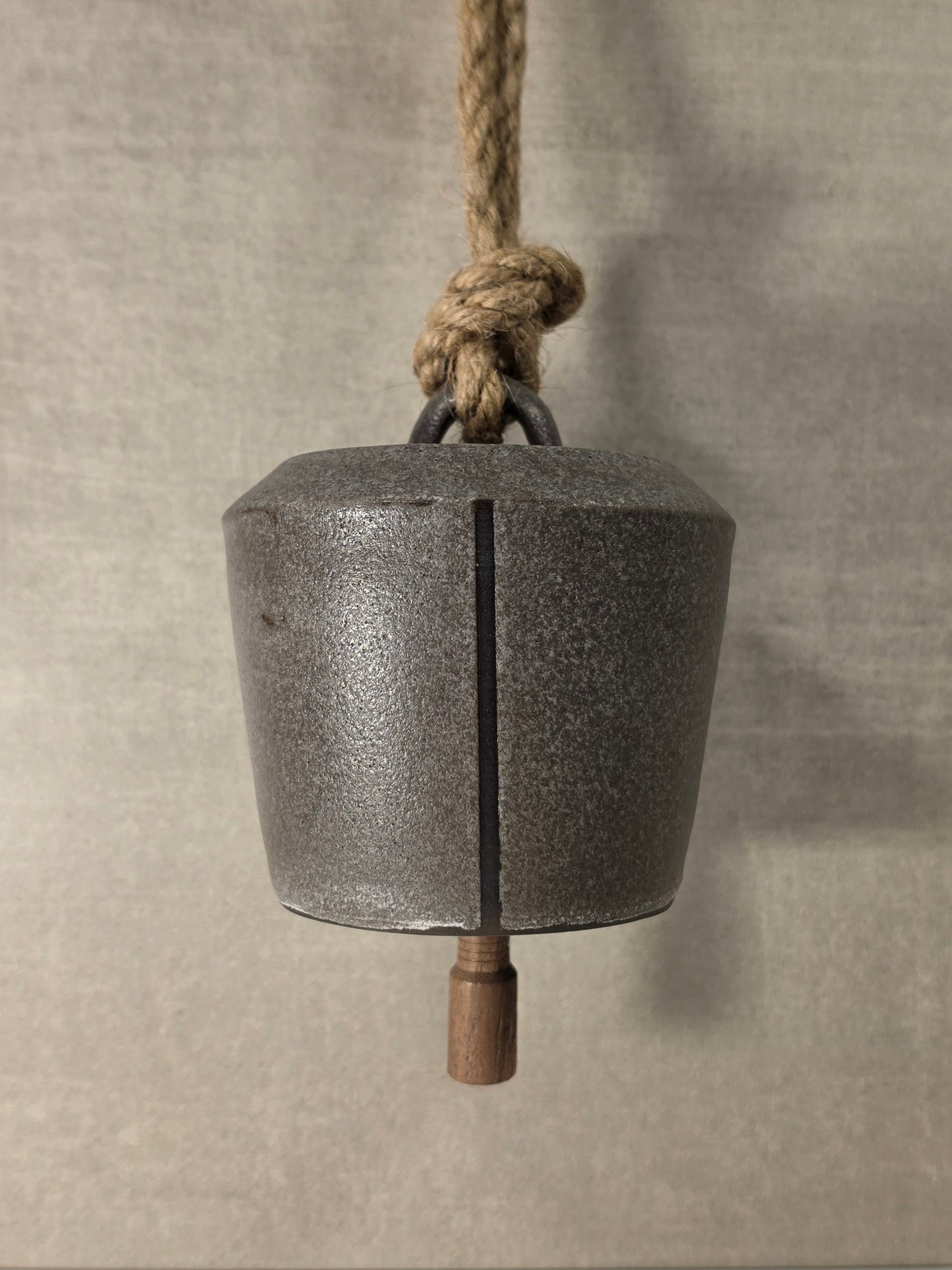 A rustic ceramic bell with a vertical stripe down the center, hanging from a thick natural fiber rope with a wooden weight at the bottom, set against a neutral textured gray background.