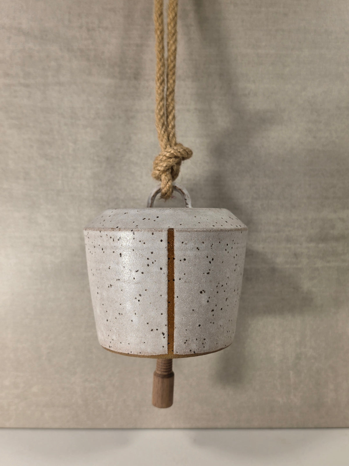 A rustic ceramic bell with a speckled off-white glaze and a vertical brown crack, hanging from a natural jute rope against a textured gray background.