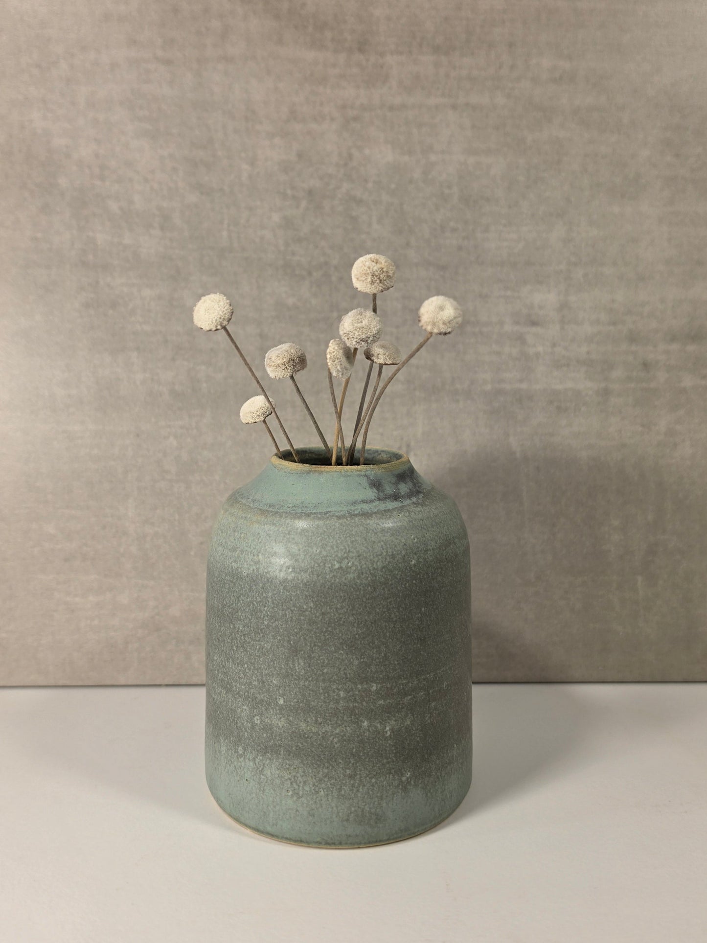A handmade ceramic vase with a textured, speckled sage green glaze, holding dried white globe flowers, set against a neutral gray textured background.