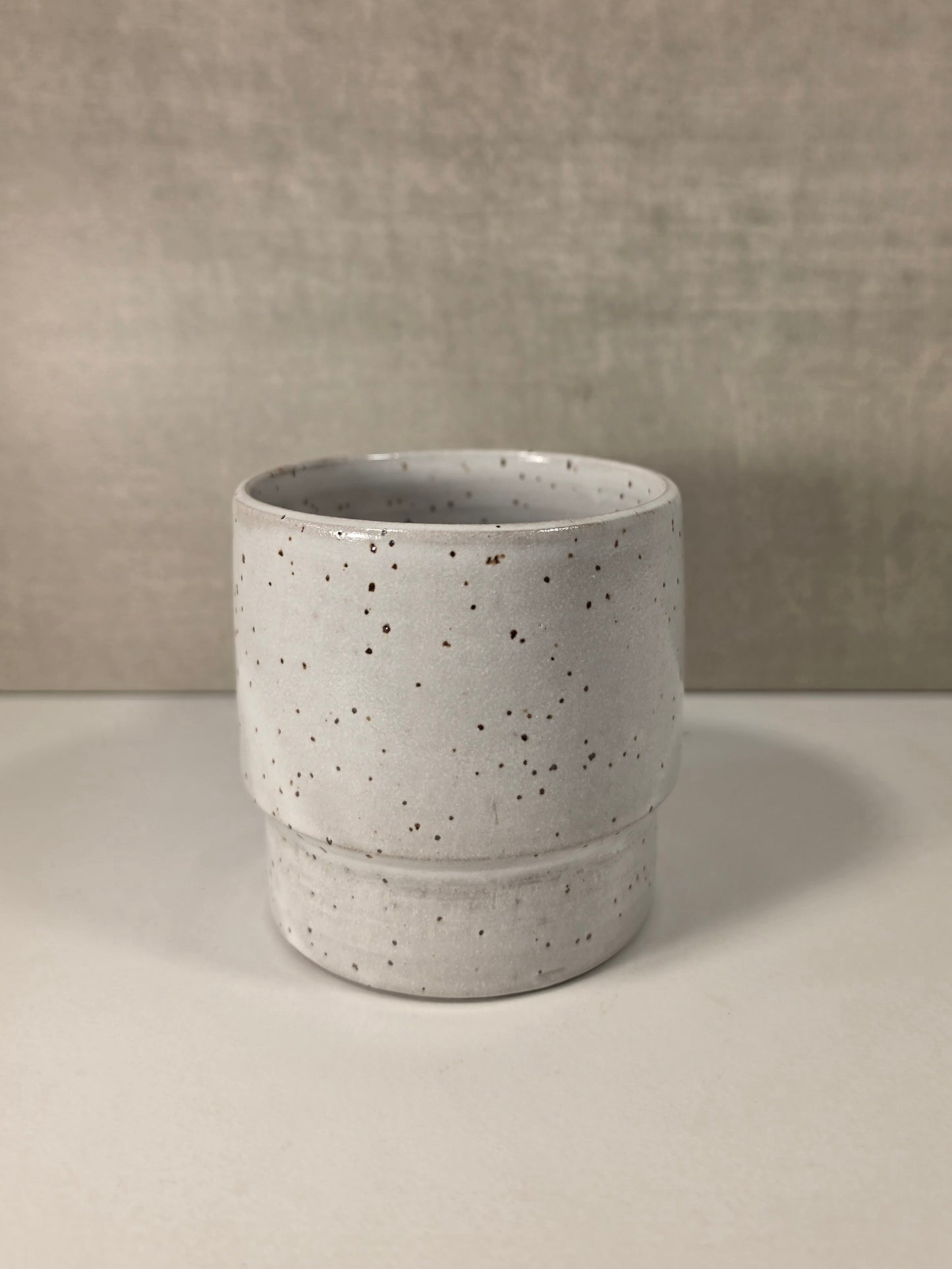 A small cylindrical ceramic cup or tumbler with a speckled off-white glaze and dark brown speckles, set against a neutral textured gray background.