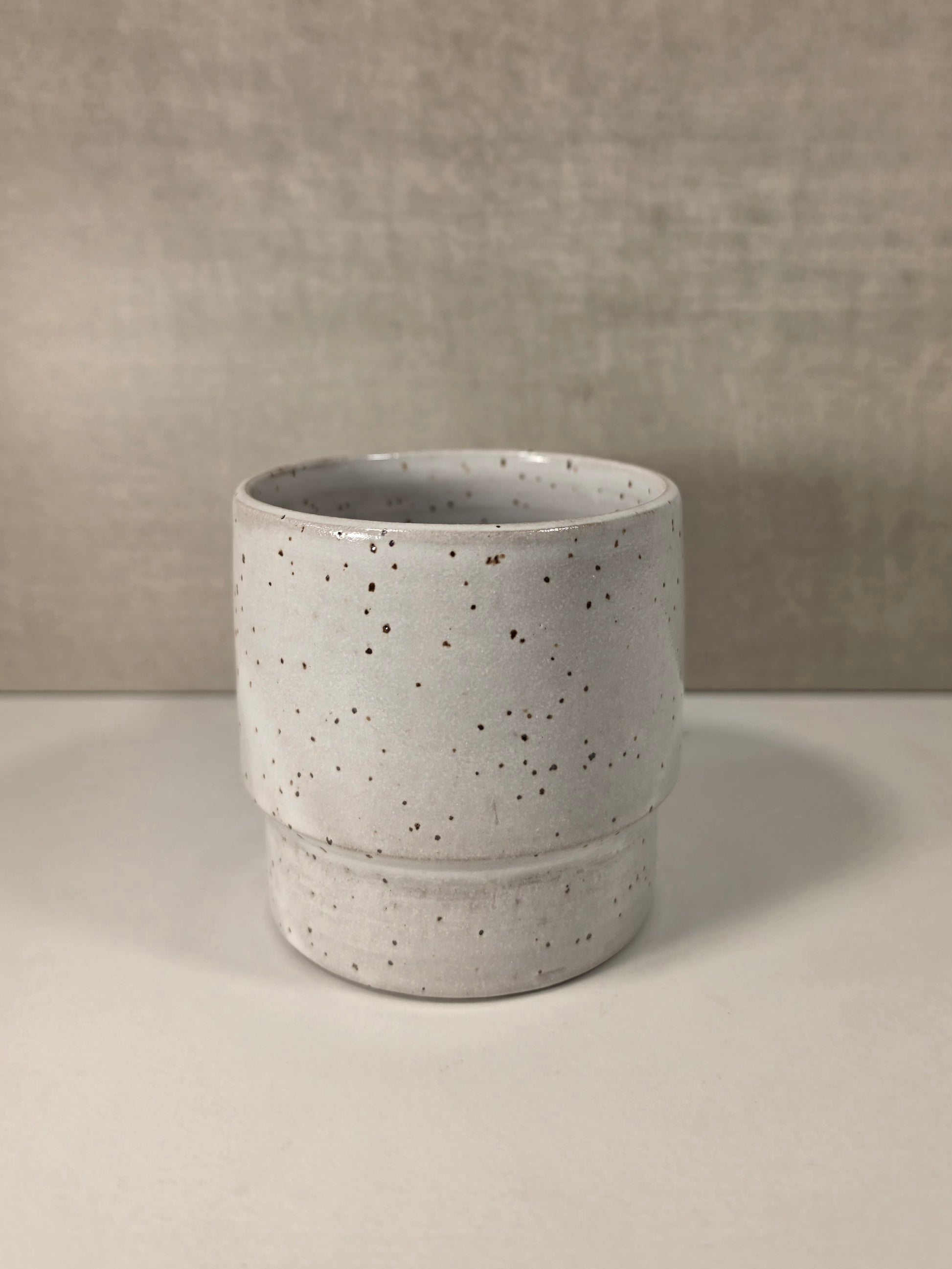 A small cylindrical ceramic cup or tumbler with a speckled off-white glaze and dark brown speckles, set against a neutral textured gray background.